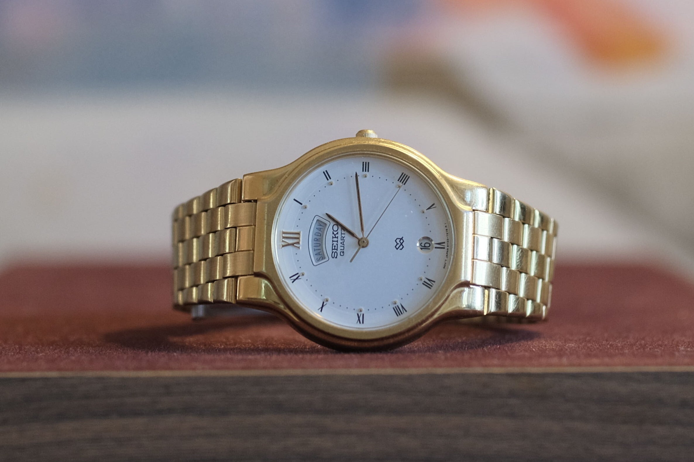 A gold-tone metal link bracelet watch with a white dial and black markers, displayed on a dark wood block pedestal with a blurred painting in the background.