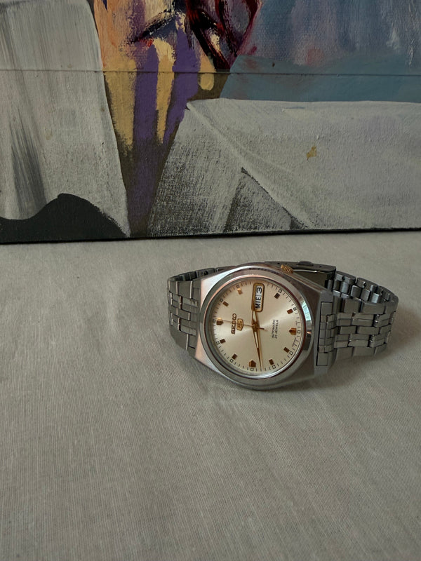 A vintage-style stainless steel wristwatch with a silver-tone metal bracelet and a cream-colored dial, resting on a textured gray surface in front of a colorful abstract painting.