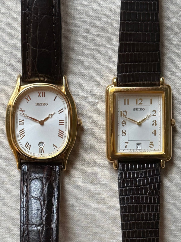 Two vintage wristwatches with gold-tone cases and white dials, one oval and one rectangular, both with dark brown textured leather straps, displayed on a light beige fabric background.
